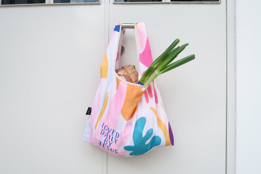 Christian gift store 
foldable reusable 
shopping bag with 
uplifting Bible verse 
"Loved Daily by Jesus" in 
vibrant pastel colors. 