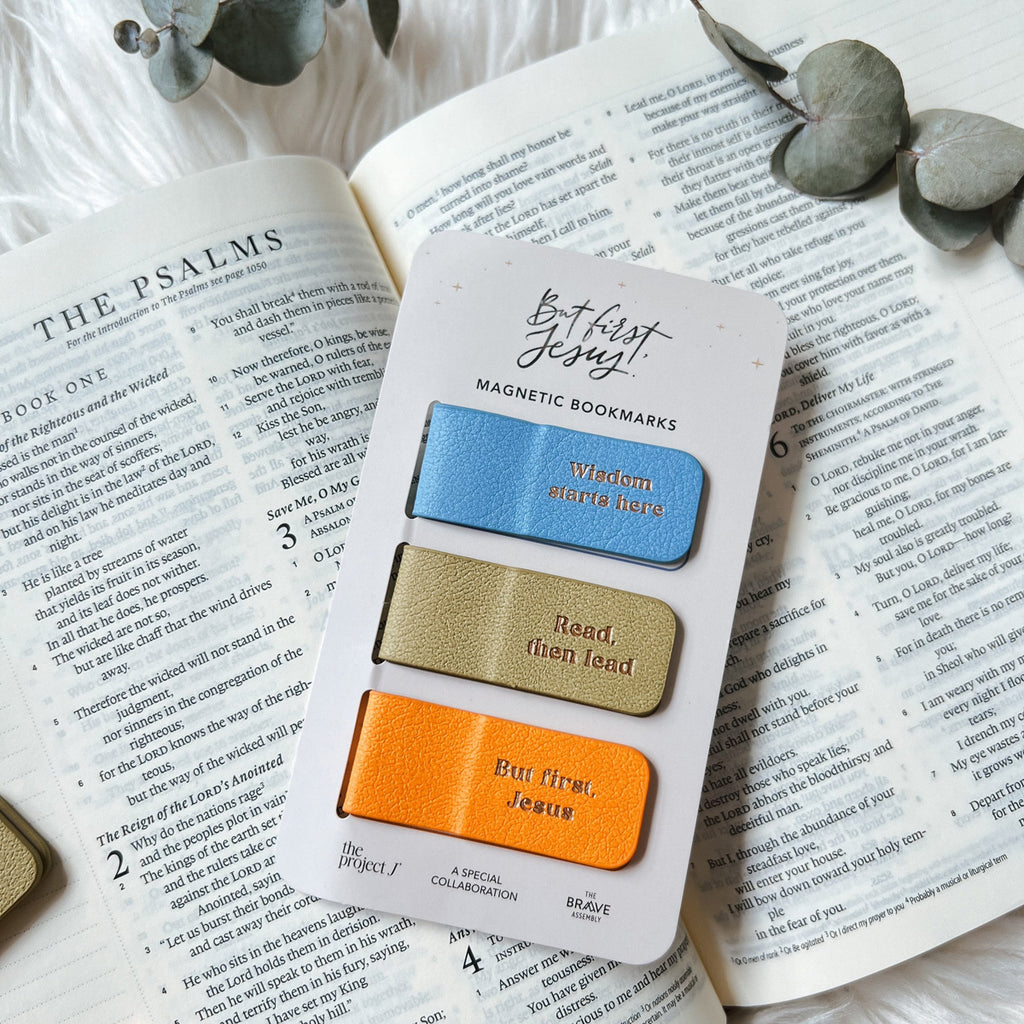 Three magnetic bookmarks in blue, olive, and orange labeled 'Wisdom starts here,' 'Read, then lead,' and 'But first, Jesus,' placed on an open Bible turned to Psalms with eucalyptus leaves on a white background.