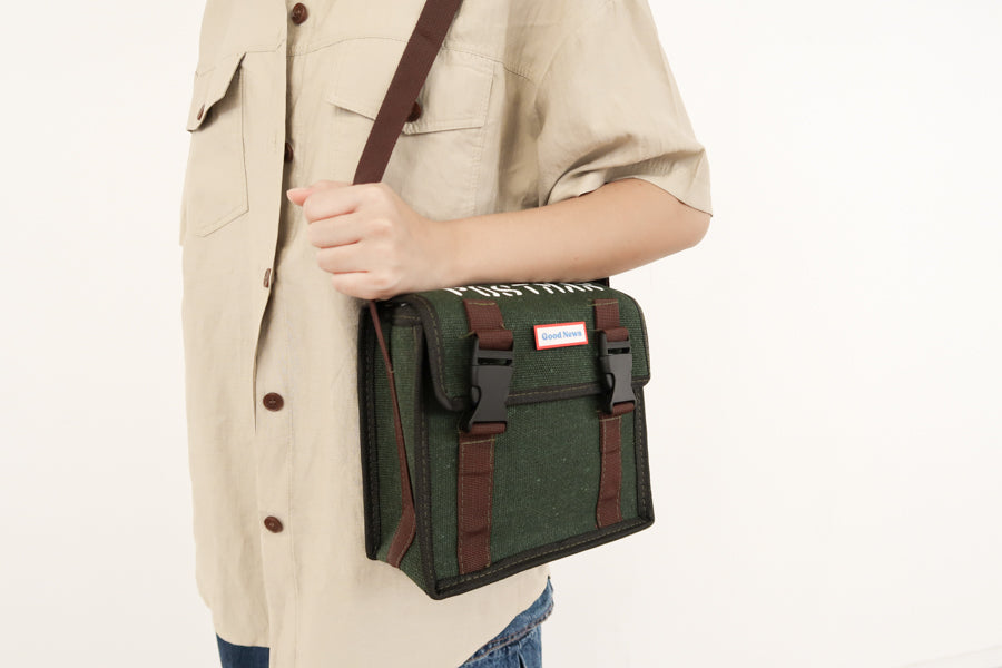 A close-up view of a person wearing a green canvas messenger bag with 'Good News' branding, highlighting the crossbody strap and faith-based design. The bag features sturdy brown straps and a durable canvas construction.