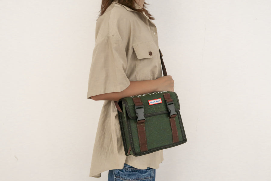 A person wearing a green canvas messenger bag with 'Good News' branding, shown from the side. The bag features brown straps and buckles, with a practical and faith-inspired design.