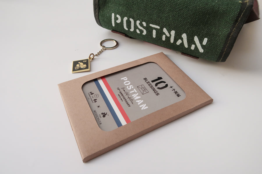 A close-up of the 'Good News' green canvas messenger bag with 'POSTMAN' text, displayed with a 10th anniversary edition 'Postman' keychain and blessings packaging. The keychain features a motorbike design, complementing the faith-inspired product set.