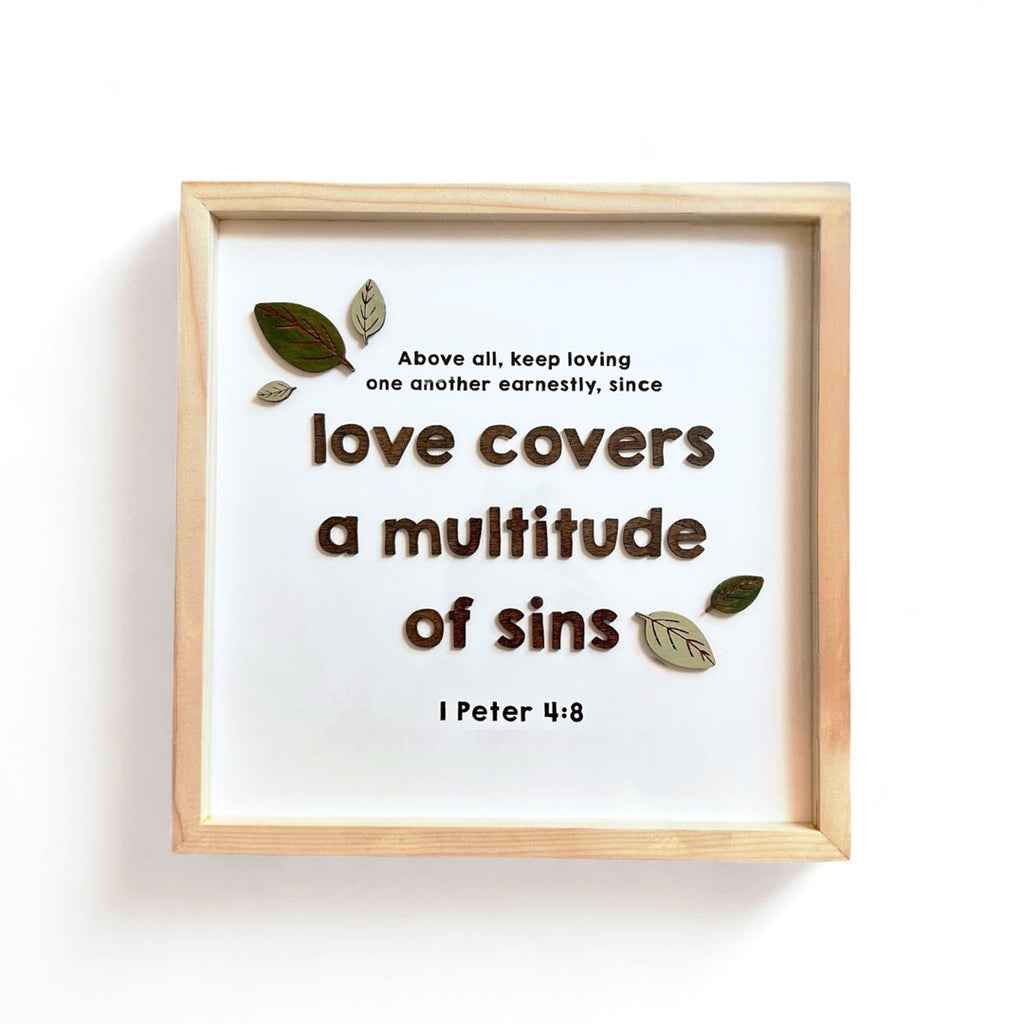 A framed Christian wall art featuring the Bible verse 'Love covers a multitude of sins' from 1 Peter 4:8. The text is in a bold, brown font with decorative leaves surrounding the quote. The minimalist wooden frame complements the simple and meaningful design.