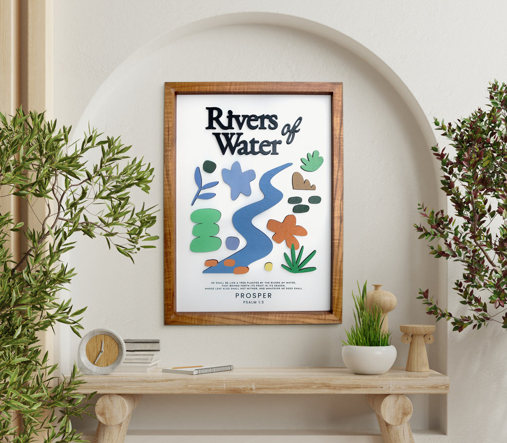 A modern wooden-framed poster featuring the Bible verse "Rivers of Water - Psalm 1:3" with colorful abstract and floral cutouts. The artwork is displayed in a bright, arch-shaped alcove surrounded by natural elements like potted plants and a wooden console table, creating a peaceful and elegant setting.