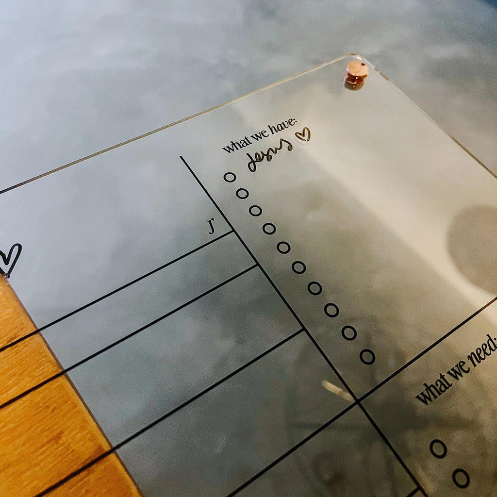 Close-up of an acrylic weekly planner board with a section labeled 'what we have' and handwritten text 'Jesus ♥', along with a task checklist on a wooden surface.