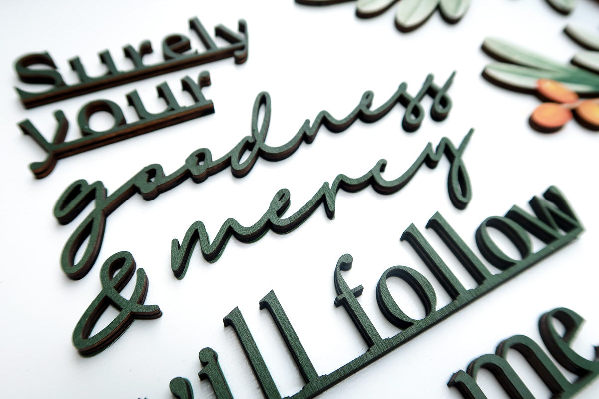 Close-up of handcrafted wooden typography reading "Surely your goodness & mercy will follow me." The layered design showcases detailed craftsmanship and elegant text on a decorative wooden poster.