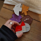 A hand holding a colorful acrylic magnet shaped like a house with the word "Home" and another magnet with the word "Grace," alongside other Christian-themed magnets on a wooden table.