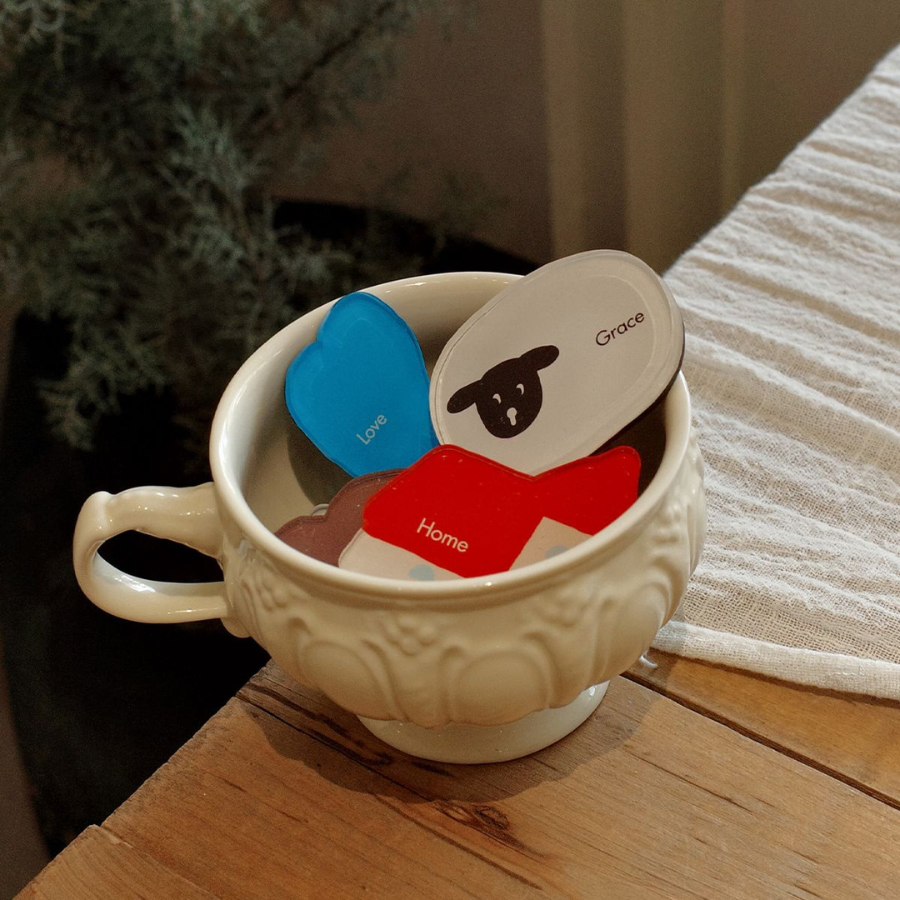 A decorative white bowl filled with colorful acrylic magnets featuring the words "Love," "Home," and "Grace," set on a wooden table with a soft cloth in the background.