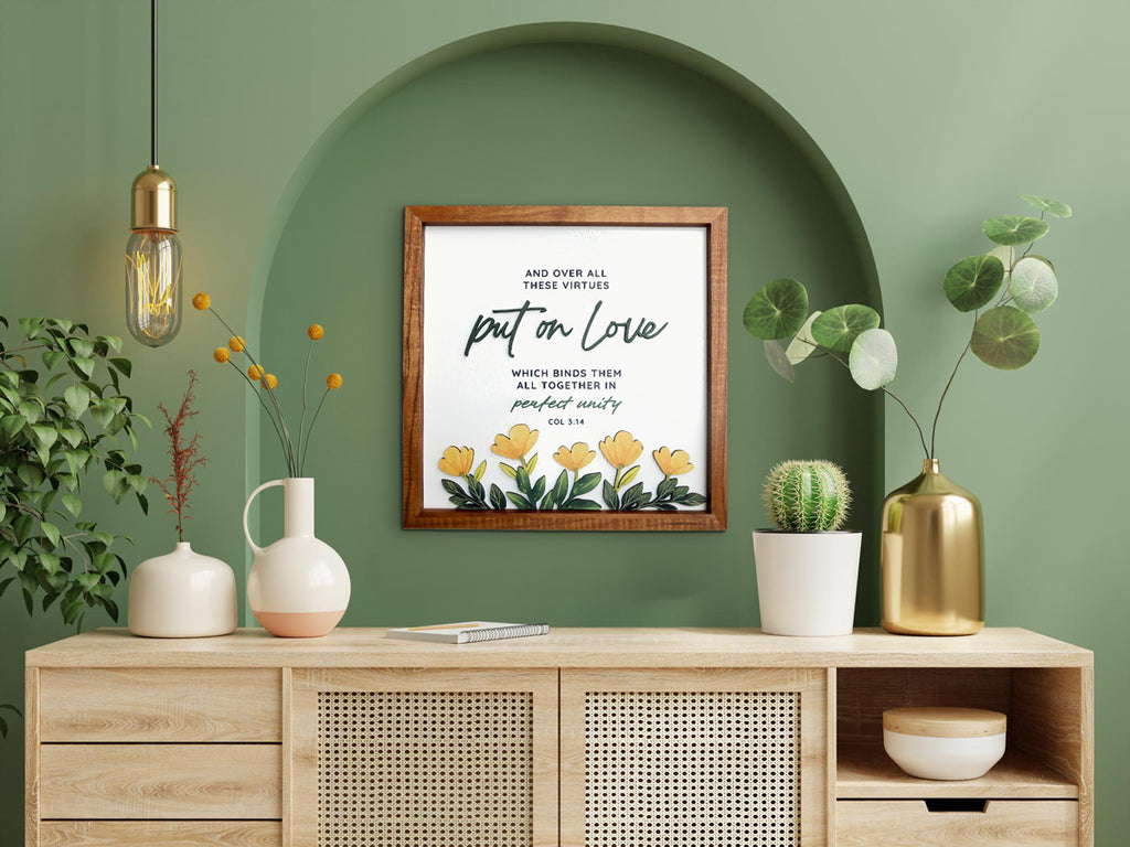 A beautifully crafted wooden-framed wall art featuring the Bible verse "Put on Love - Colossians 3:14." The poster includes yellow floral designs and an elegant script style. Displayed in a modern setting with green walls, an arched alcove, and decor elements like a wooden console, potted plants, and warm lighting, creating a serene and sophisticated ambiance.