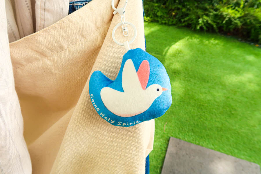 Blue plush keychain featuring a white dove and the phrase 'Come Holy Spirit,' attached to a beige bag as a symbol of faith and peace.