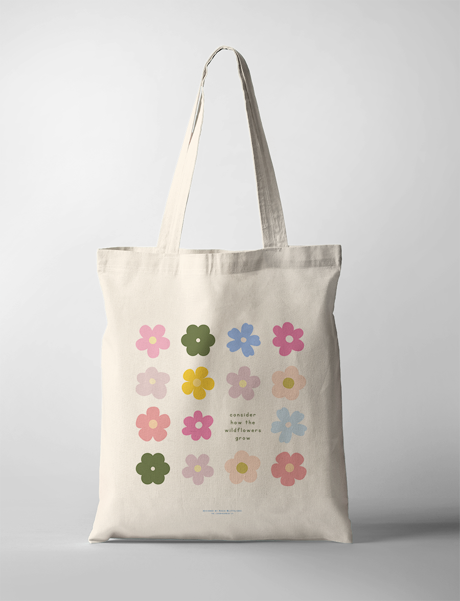 Neutral canvas tote bag decorated with colorful wildflower illustrations and the uplifting phrase "consider how the wildflowers grow," embodying simple happiness and faith-inspired encouragement.