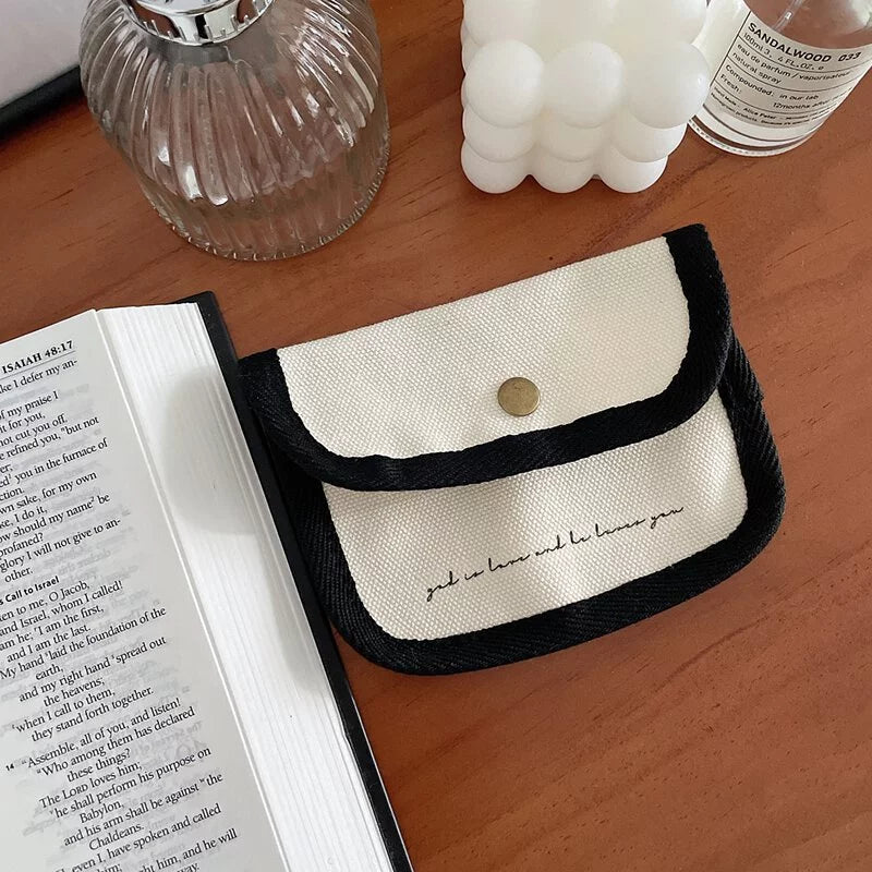 Cream canvas coin purse with black trim and a snap closure, placed on a wooden desk beside an open Bible and decorative items