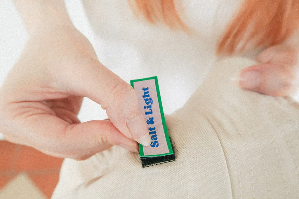 A close-up of a hand attaching a green and white velcro patch labeled "Salt & Light" onto a beige cap, showcasing customizable and interchangeable faith-based designs.