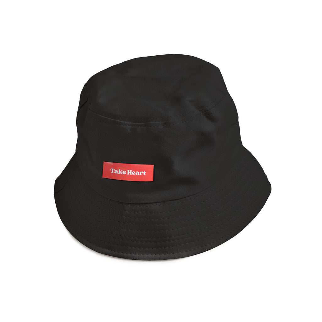 A black bucket hat featuring a red patch with white text reading "Take Heart," a Christian streetwear accessory promoting courage and faith.