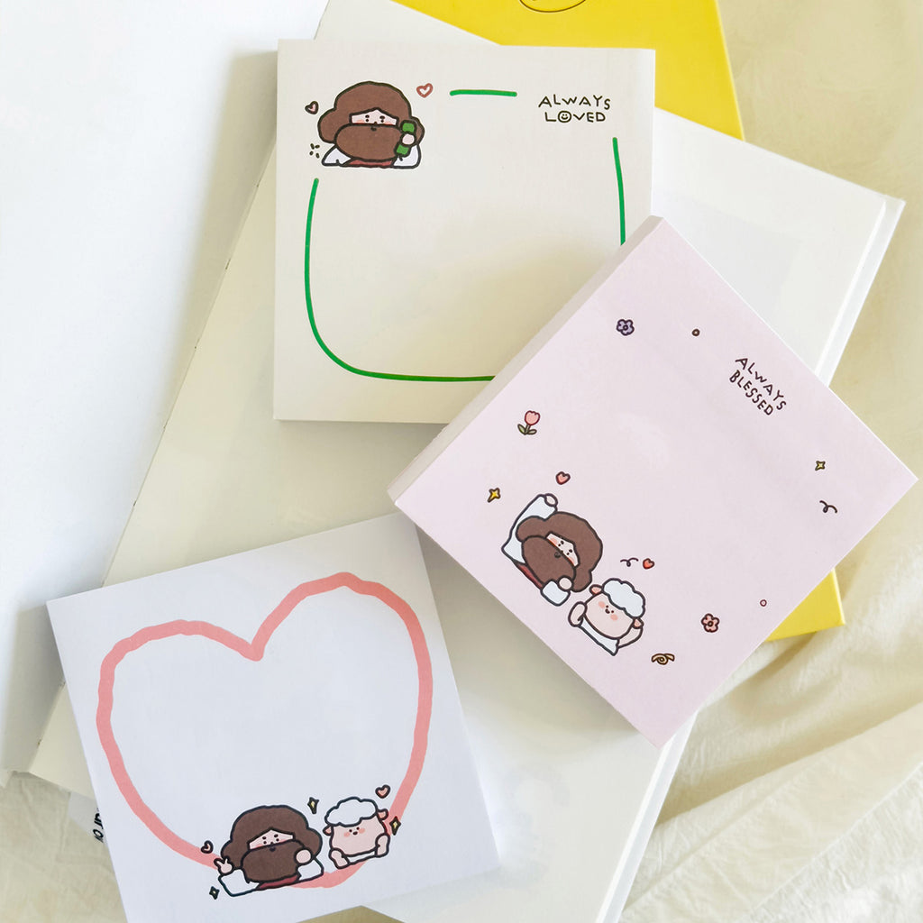 A set of three Christian-themed sticky notes, including designs with Jesus and a sheep, a pink heart, and phrases like 'Always Loved' and 'Always Blessed,' laid on a stack of books.