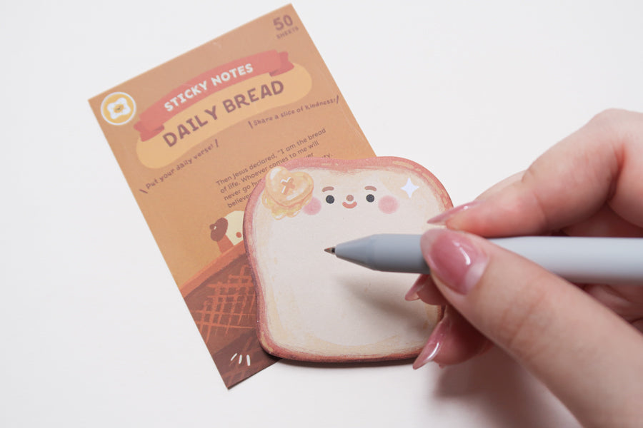 Close-up of Daily Bread bread-shaped sticky note being written on, Christian notepad for journaling and reminders.