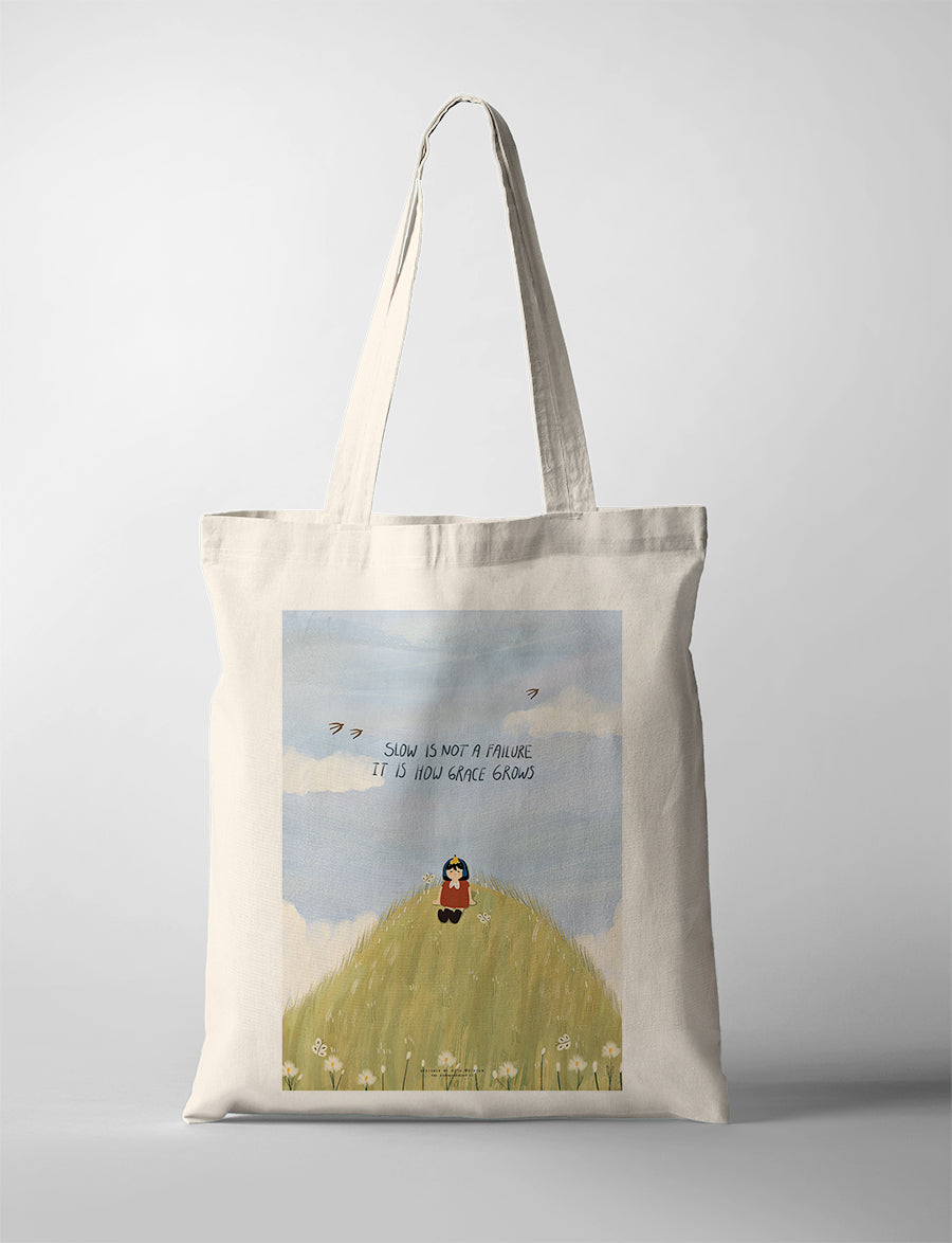 Canvas tote bag with Dita illustration of a girl sitting on a grassy hill with the phrase “Slow is not a failure, it is how grace grows” against a soft sky background