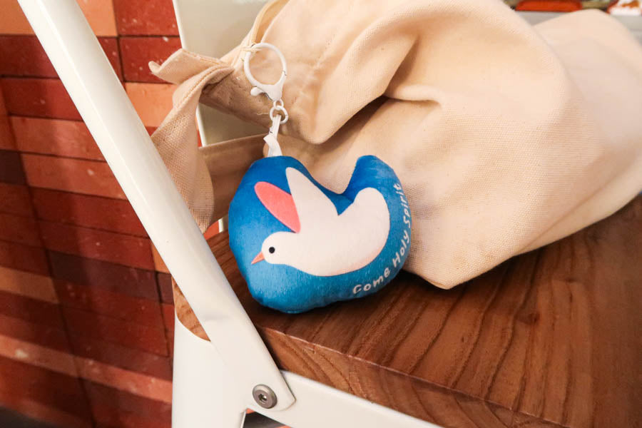 A blue plush keychain with a white dove illustration and the phrase 'Come Holy Spirit,' symbolizing peace and faith in a compact accessory