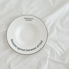 White ceramic plate with the inspirational messages "God loves you every day" and "Dreams sprout because of God," placed on a soft white fabric, encouraging faith and simple happiness.