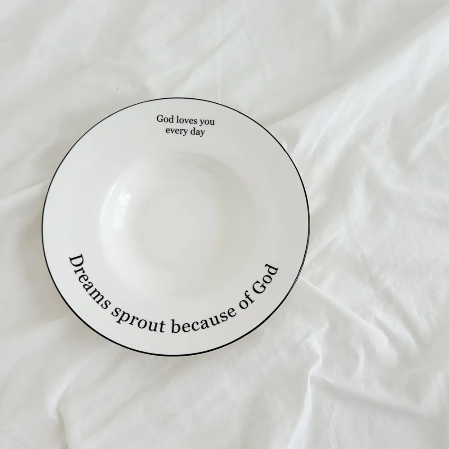 White ceramic plate with the inspirational messages "God loves you every day" and "Dreams sprout because of God," placed on a soft white fabric, encouraging faith and simple happiness.