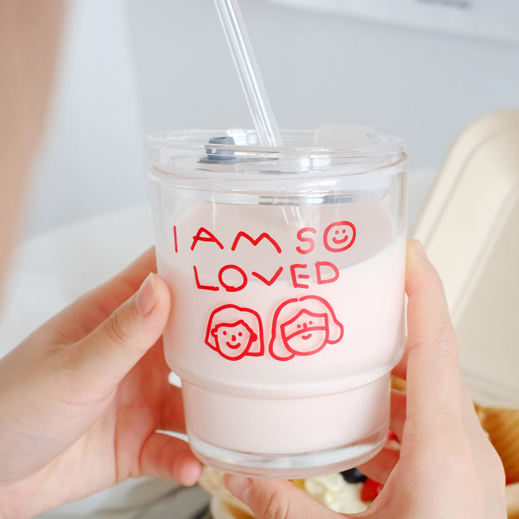 Hands holding a clear glass tumbler filled with pink milk, featuring red text saying 'I Am So Loved' with smiley face illustrations and a reusable straw.