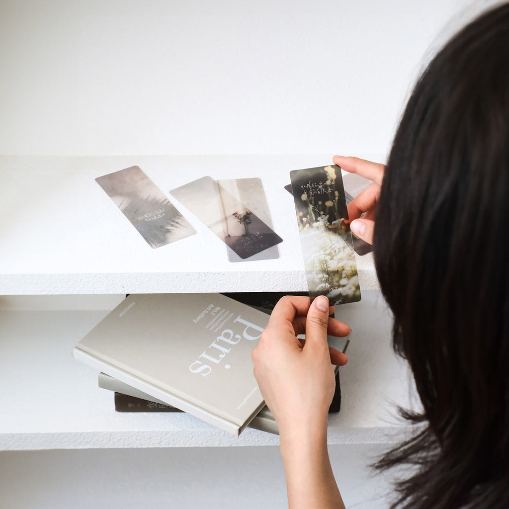 A woman selecting a minimalist, nature-inspired bookmark from a collection, featuring faith-based designs and soft, elegant tones, perfect for gifts and stationery enthusiasts.