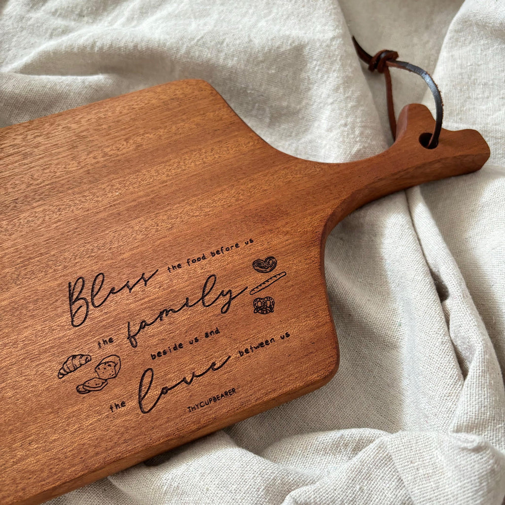 A beautifully engraved wooden cheese board featuring the text "Bless the food before us, the family beside us, and the love between us." The rustic board design includes small engraved illustrations of bread and hearts, making it a perfect kitchen accessory or serving piece. Styled on a soft neutral fabric for a cozy and elegant presentation.