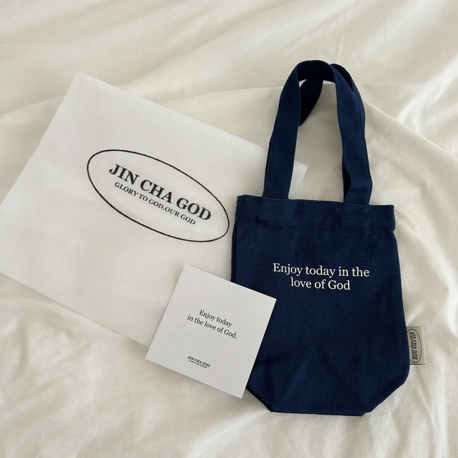 Navy blue beverage bag with the message "Enjoy today in the love of God," placed beside an open Bible and a cozy bedside table, inspiring faith and simple happiness.