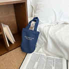 Navy blue beverage bag with the uplifting message "Enjoy today in the love of God," placed on a wooden table beside a framed Bible verse, inspiring simple happiness and faith.