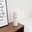  Cream thermos bottle with the words "enjoy today in the love of God," placed on a wooden bedside table in a cozy, minimalist room, inspiring daily encouragement and simple happiness.