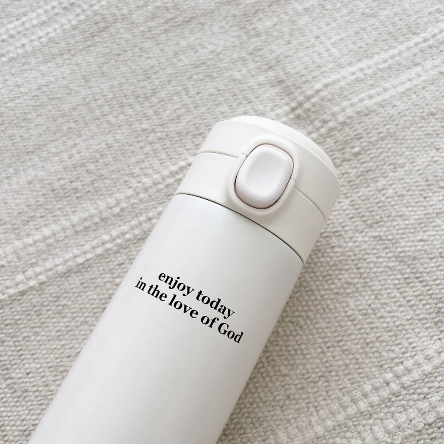 Close-up of a cream thermos bottle with the words "enjoy today in the love of God," resting on a textured fabric, offering daily encouragement and simple happiness.