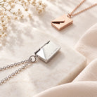 Silver and rose gold envelope locket necklaces styled on a light stone surface