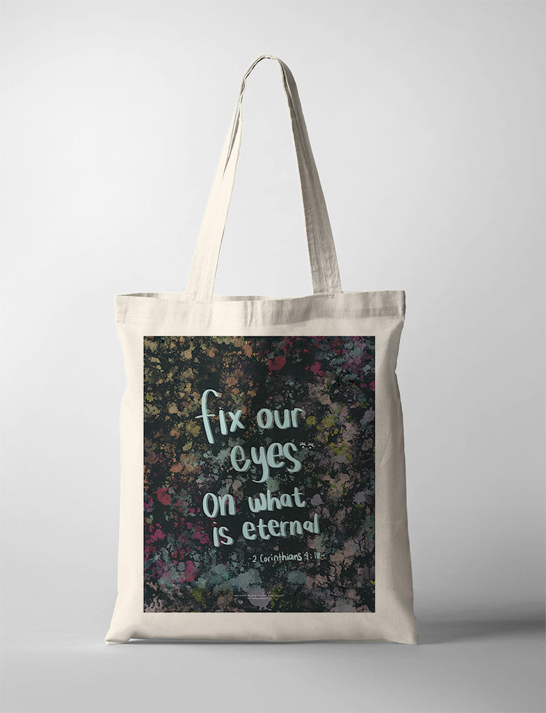 A beige tote bag featuring a design with the Bible verse "Fix our eyes on what is eternal" from 2 Corinthians 4:18, displayed in handwritten-style text over a vibrant, abstract background.