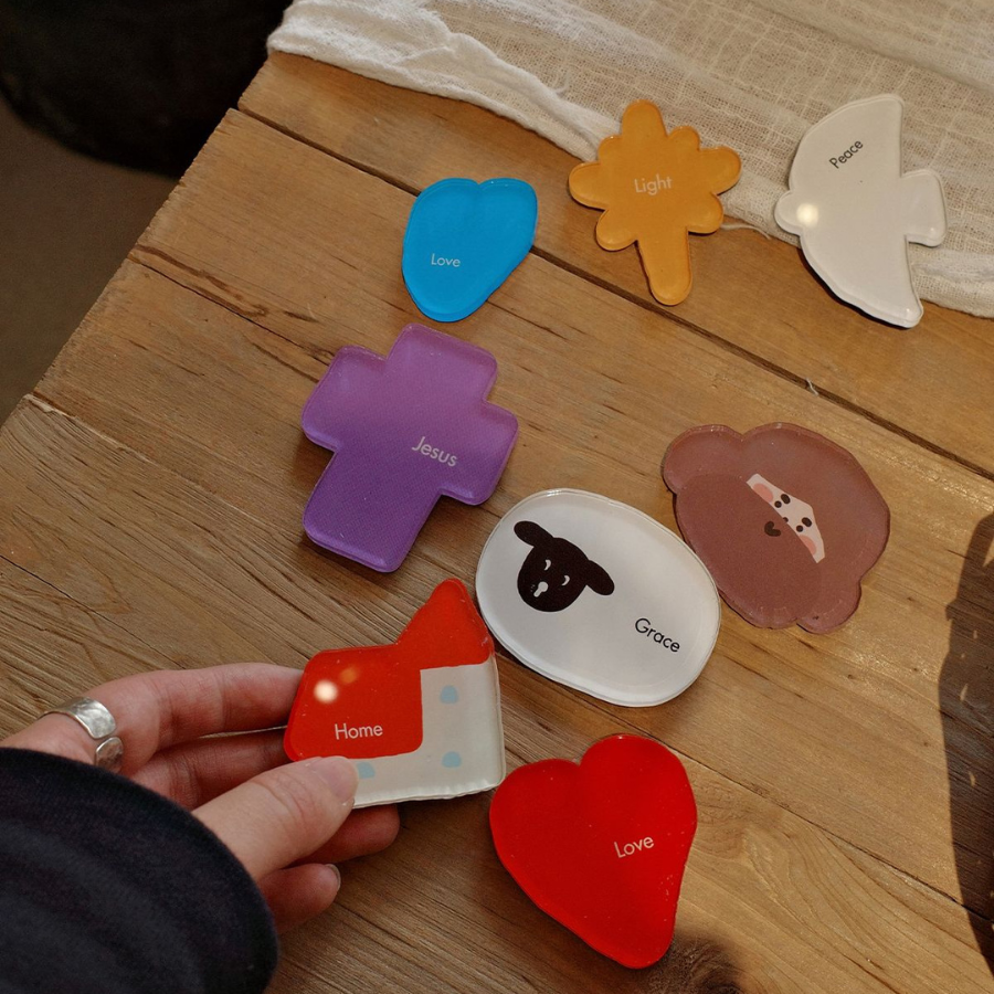 A hand holding colorful acrylic magnets with Christian symbols and words like "Love," "Jesus," and "Grace" on a wooden table.