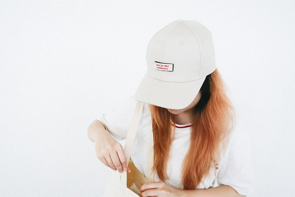 A person with long, orange-tinted hair wearing a beige cap featuring a red and white velcro patch that says "Joy in the ordinary," highlighting a customizable faith-inspired accessory.