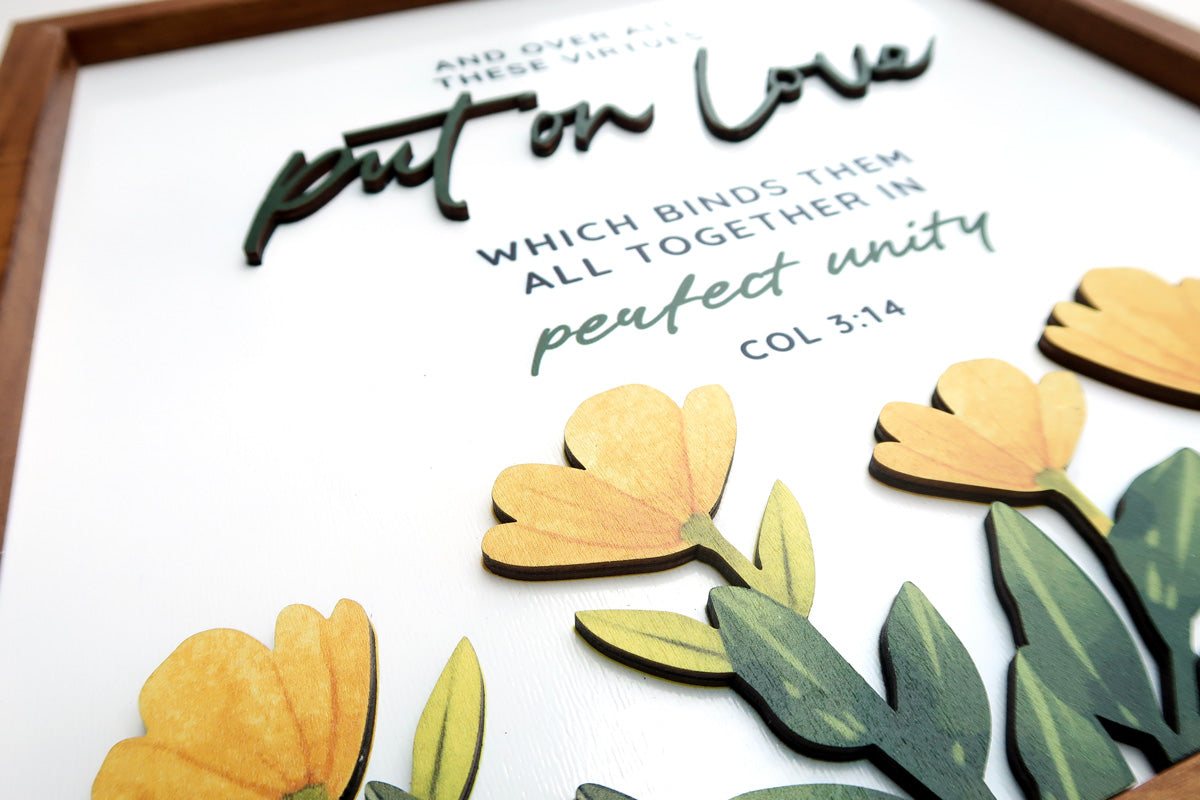 Close-up of a wooden scripture poster featuring the text "Put on Love" from Colossians 3:14. Includes layered lettering and decorative yellow flowers with green leaves.