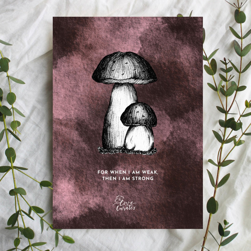 A greeting card with a black and white illustration of two mushrooms on a textured dark pink background. The card features the quote 'For when I am weak, then I am strong,' conveying a message of strength through vulnerability. Green foliage decorates the sides of the card.