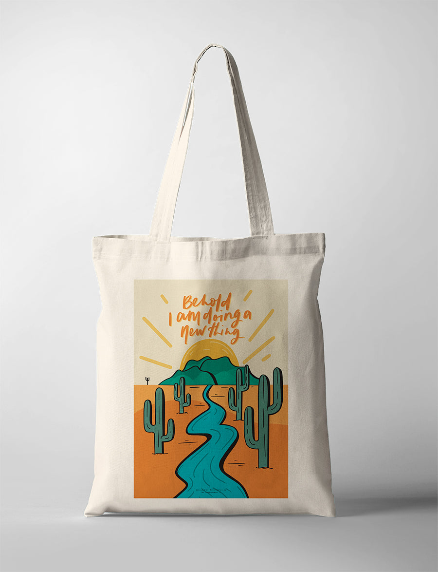Christian canvas tote bag featuring “Behold I am doing a new thing” Bible verse with illustrated desert landscape, river, and sunrise, designed by Fouronetwo.sg in collaboration with The Commandment Co.
