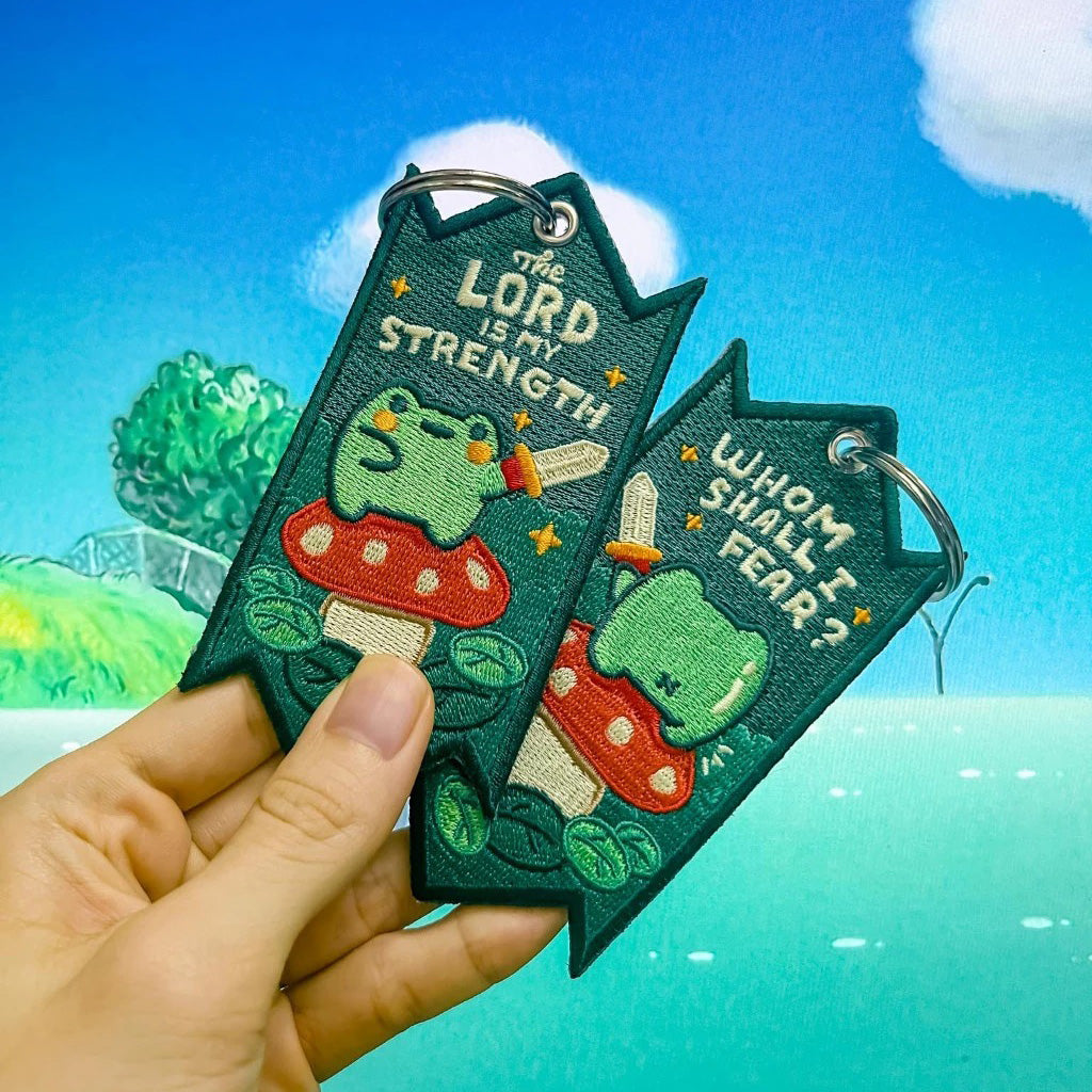 Two Christian-themed embroidered keychains featuring a green frog on a red mushroom with swords, displaying the messages 'The Lord is My Strength' and 'Whom Shall I Fear?' held against a blue sky background.