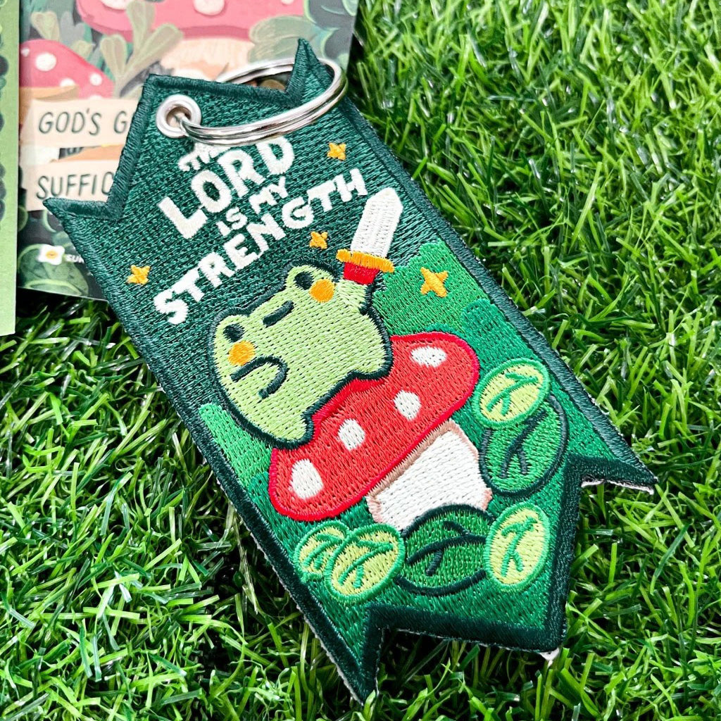 Christian embroidered keychain featuring a green frog on a red mushroom holding a sword, with the message 'The Lord is My Strength,' set on vibrant green grass.