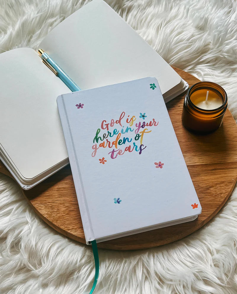 Off-white hard cover journal with “God is here in your garden of tears” text design by Project J placed on wooden tray beside open notebook and candle.