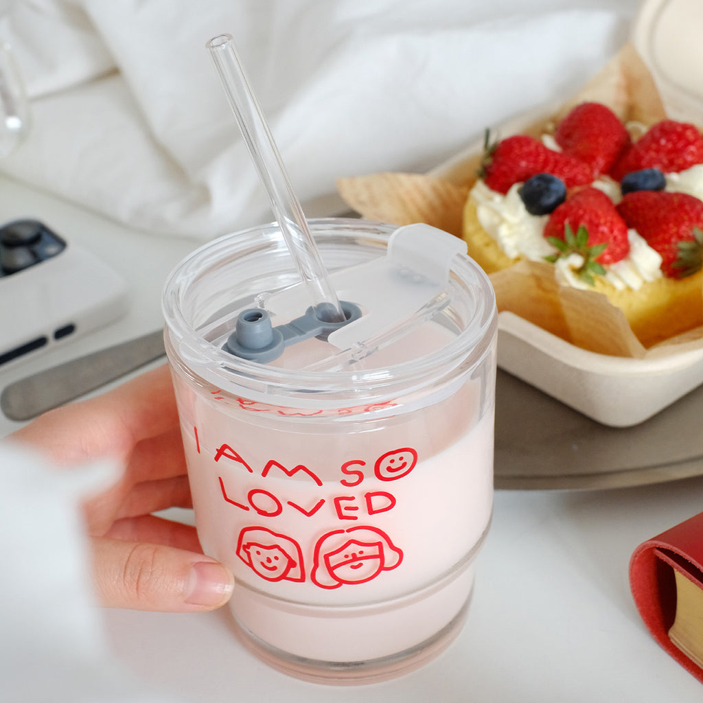 A clear glass tumbler with red 'I Am So Loved' text, reusable straw, and pink milk, placed next to a dessert topped with fresh strawberries and blueberries.