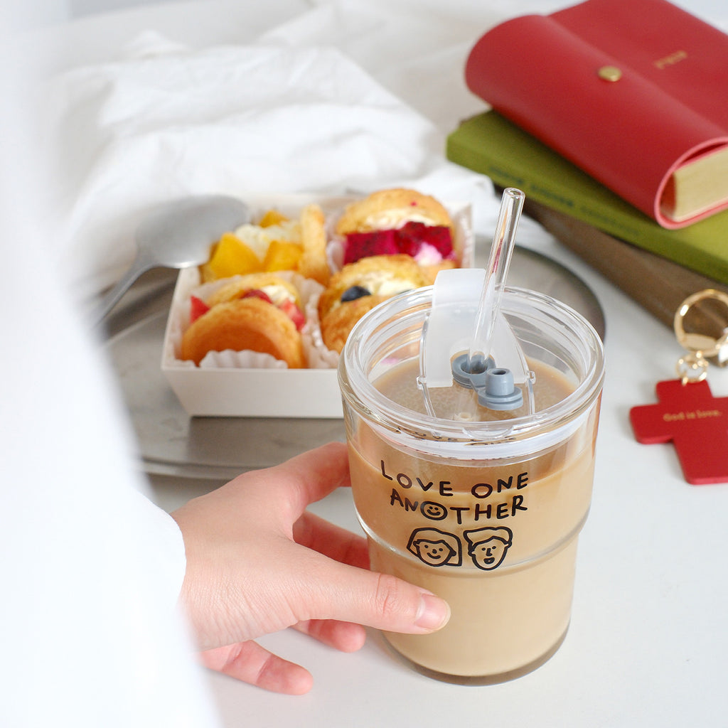 A clear glass tumbler featuring the 'Love One Another' design, filled with coffee and paired with a dessert box and books in the background.