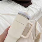 Hand holding a cream insulated cup with the words "God has you in His heart," resting on a cozy white bed, offering encouragement and simple happiness as a Christian gift from Jincha God