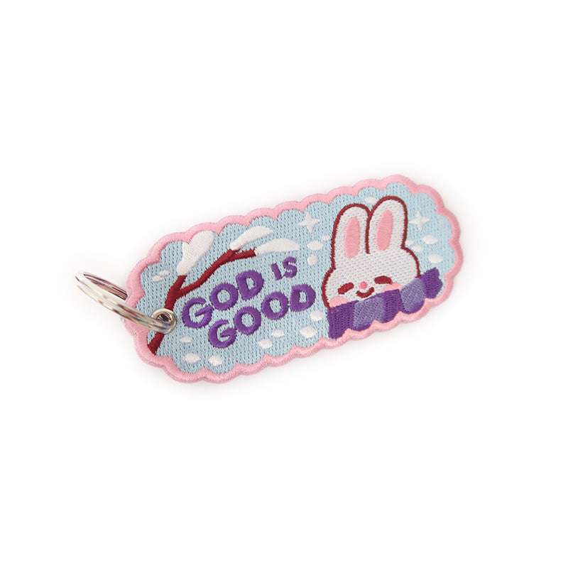 Smiling white bunny on a blue keychain with the phrase 'GOD IS GOOD' in embroidery.