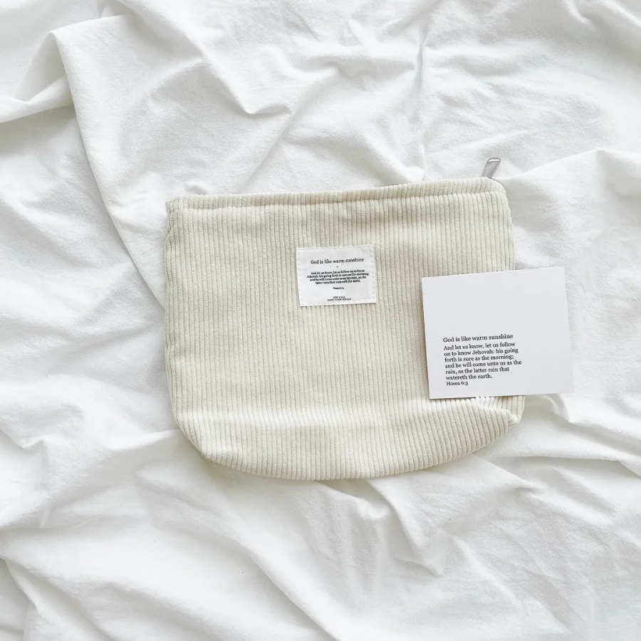 God is like warm sunshine zipper clutch in cream corduroy with an inspirational message patch, displayed on a soft white fabric, inspiring simple happiness and faith.