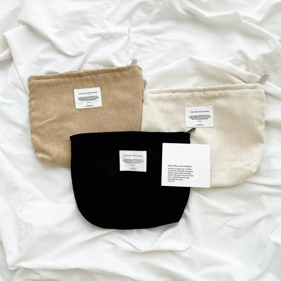 God is like warm sunshine zipper clutch in beige, cream, and black corduroy, each with an uplifting message patch, displayed on a cozy white fabric to inspire simple happiness and faith.