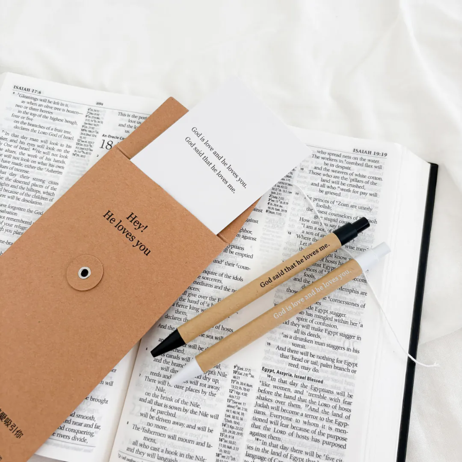 God is Love Pen Set with Bookmark placed on an open Bible, featuring uplifting messages and kraft packaging that reads "Hey! He loves you."