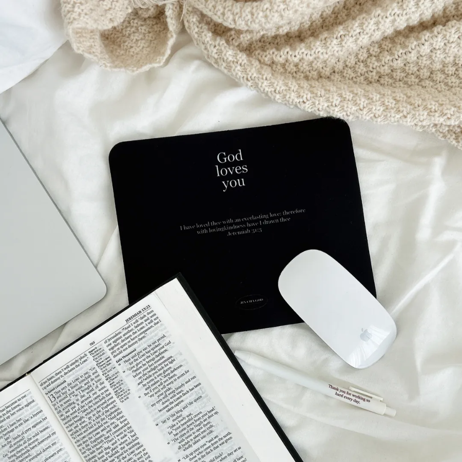 Black mouse pad with the words "God loves you" and Jeremiah 31:3, placed on a cozy bed beside a Bible, pen, and laptop, offering daily encouragement and simple happiness.