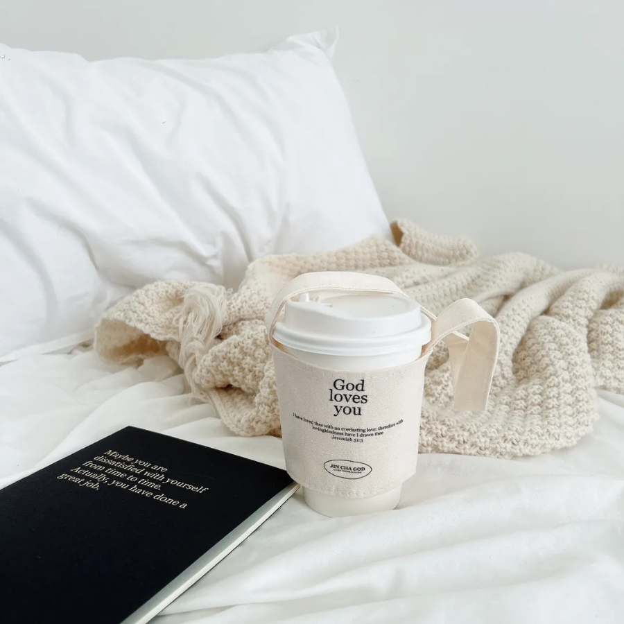 Reusable cup holder with "God loves you" and Jeremiah 31:3 Bible verse, displayed on a cozy bed with a knitted blanket and inspirational notebook, perfect Christian eco-friendly gift.