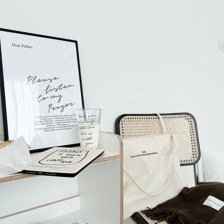 God Your love is my dependence canvas tote bag on a rattan chair, beside a cozy desk with an inspirational prayer print, a faith-themed glass, and a soft brown scarf, creating a peaceful and uplifting atmosphere.
